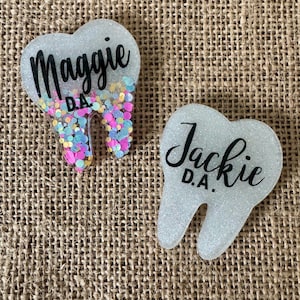 May include: Two tooth-shaped name badges. One badge has the name "Maggie D.A." in black script with colorful confetti at the bottom. The other badge has the name "Jackie D.A." in black script. Both badges are white with glitter.