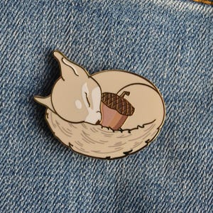May include: A gold-rimmed enamel pin featuring a cream-coloured fox curled into a circle while sleeping. The fox has closed eyes and is holding a brown acorn with a textured cap against its body. The design is outlined in thin gold metal lines.