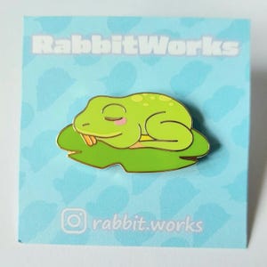 May include: A green enamel pin depicting a sleeping frog on a lily pad. The frog has closed eyes and pink cheeks. The pin is on a light blue card with the text "RabbitWorks" and the Instagram handle "rabbit.works".