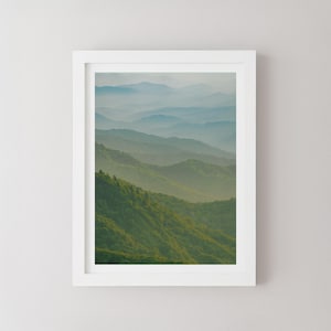 Puede incluir: Una impresión enmarcada de un paisaje montañoso. La imagen muestra una serie de colinas cubiertas de árboles verdes, que se desvanecen en un tono azul brumoso en la distancia. La obra de arte se presenta dentro de un marco blanco.