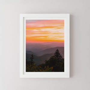 May include: A framed landscape print featuring a mountain range at sunset. The sky transitions from orange to pink, with layers of blue and purple mountains in the distance. The print is in a white frame.