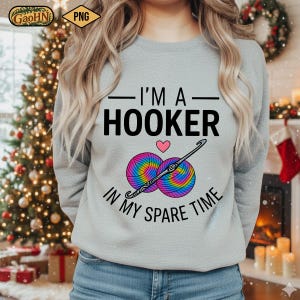 May include: A light gray sweatshirt with the text "I'M A HOOKER IN MY SPARE TIME" in black. Below the text are two rainbow-colored yarn balls and a crochet hook. A pink heart is between the text and the yarn.