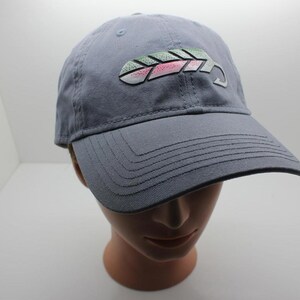 Puede incluir: Gorra de béisbol gris con visera curva. La gorra presenta un diseño bordado de un pez estilizado con detalles verdes, rosas y blancos. La gorra se muestra en una cabeza de maniquí.