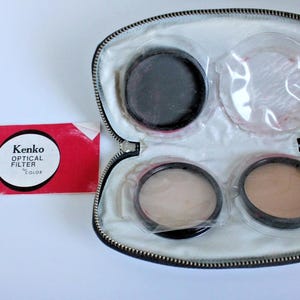 May include: A vintage Kenko Optical Filter set in a zippered case. The case is open, revealing four circular filters and a red card with the Kenko logo. The filters are various shades of black, beige, and clear.