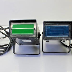 May include: Two black stage lights with rectangular green and blue lenses. Each light has a black stand and a black power cord. The lights are on a white surface.