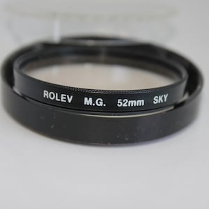 May include: A black camera lens filter with the text "ROLEV M.G. 52mm SKY" in white. The filter is circular and appears to be made of glass, with a black metal ring. The filter is designed to reduce the amount of blue light.