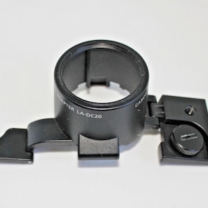May include: Black Canon LA-DC20 adapter. The adapter has a circular opening and a rectangular base with a screw. The text "ADAPTER LA-DC20" is visible on the side. The adapter is designed for camera use.