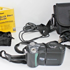 Könnte beinhalten: Eine schwarze Nikon Coolpix 4500 Digitalkamera mit Zubehör, darunter ein Objektiv, eine Tragetasche, ein Ladegerät und eine Kurzanleitung. Die Kamera hat einen schwarzen Gurt und einen grünen Akzent. Die Anleitung ist gelb mit schwarzem Text.