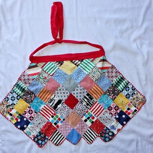 May include: A patchwork apron with a red neck strap and trim. The apron is made of various patterned squares in colors like red, blue, yellow, and green. The apron has a scalloped edge.