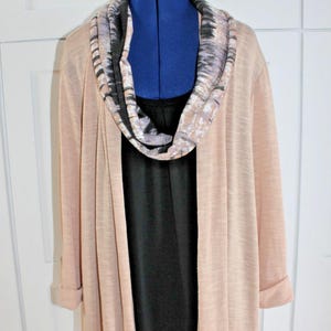 May include: A light peach-colored long cardigan with rolled-up sleeves is draped over a black top. A patterned cowl neck scarf in shades of gray, black, and beige adds a stylish accent. The outfit is displayed on a blue dress form.
