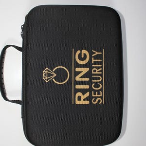 以下が含まれることがあります： 黒い長方形のケースで、ハンドルとジッパーが付いています。ケースには、金のダイヤモンドリングのグラフィックと、金色の文字で「RING SECURITY」という文字が描かれています。このケースは、ジュエリーを保護するように設計されています。