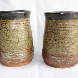 May include: Two handmade ceramic beer steins. The mugs have a speckled tan and brown glaze with a dark brown rim and a reddish-brown base. Each mug has a unique, curved handle.