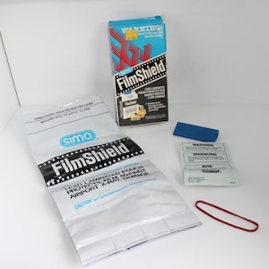 May include: A collection of vintage FilmShield products. Includes a blue and red box, a silver pouch with black text, a blue plastic piece, a small paper with text, and a red rubber band. The box reads "FilmShield".