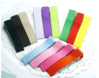 May include: A collection of colorful ribbon-covered barrettes in various shades, including black, brown, beige, white, purple, green, red, yellow, light blue, pink, and orange. The barrettes are arranged on a white, lacy surface.