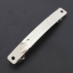 May include: A silver-colored metal hair barrette with a rectangular shape. The barrette has a decorative design in the center and a small hole at each end. The clasp is visible on the underside.