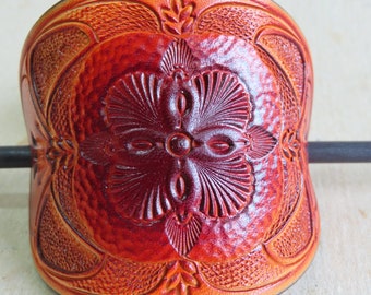 May include: A handcrafted, reddish-brown leather hair barrette with an embossed floral design. The barrette features a central flower motif surrounded by intricate leaf patterns and textured details. A dark metal stick is inserted through the barrette.