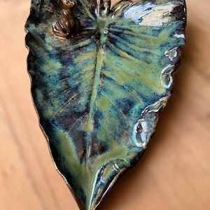 Può includere: Un piatto in ceramica a forma di foglia con una piccola statuina di gatto. Il piatto presenta una finitura lucida con sfumature di verde e blu e un bordo color bronzo. Il gatto è color bronzo.