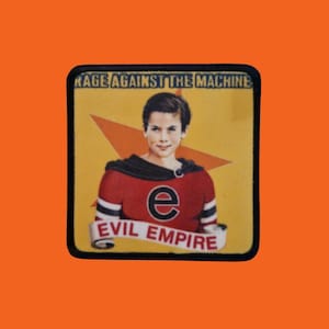 Può includere: Toppa quadrata con una persona in un top rosso e nero, la lettera "e" e le parole "EVIL EMPIRE" su uno striscione. La toppa ha il testo "RAGE AGAINST THE MACHINE" in alto e uno sfondo giallo con una stella rossa.