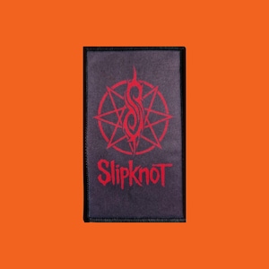 Pode incluir: Um patch retangular com o nome da banda "Slipknot" em letras góticas vermelhas. Acima do nome, um símbolo vermelho dentro de um desenho de estrela, sobre um fundo cinza escuro com uma borda preta. Para roupas ou acessórios.