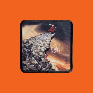 Pode incluir: Um patch quadrado com o logotipo da banda Korn e a arte do álbum. A imagem mostra uma figura em vermelho no topo de um penhasco, com as palavras "Korn" e "Follow the Leader". O patch tem uma borda preta e um fundo laranja.
