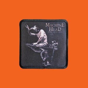 Op de afbeelding: Vierkante zwarte patch met een zwarte rand. De patch heeft de tekst "MACHINE HEAD" in het wit, boven een zwart-wit illustratie van een figuur. De figuur zit op een donkere ondergrond, met een donkere achtergrond.