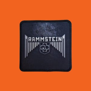 Puede incluir: Un parche cuadrado negro con el nombre de la banda "RAMMSTEIN" en letras blancas. Debajo, un emblema blanco con un escudo y un símbolo en forma de cruz, flanqueado por líneas verticales.