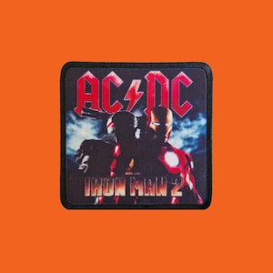 May include: Square patch with the AC/DC logo in red with a lightning bolt, and the words "IRON MAN 2". The patch has a black border and an image of Iron Man. The background is a mix of blue and white.