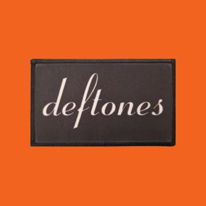 Könnte beinhalten: Ein rechteckiger schwarzer Aufnäher mit dem Wort "deftones" in weißer Schreibschrift. Der Aufnäher hat einen schwarzen Rand und ist vor einem orangefarbenen Hintergrund platziert.