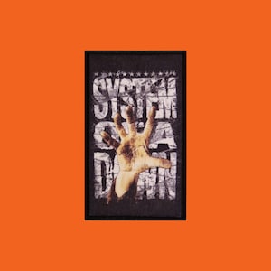 Peut inclure: Un écusson noir avec le nom du groupe "SYSTEM OF A DOWN" en blanc, avec un effet vieilli. Une main détaillée, de couleur beige, tend la main depuis le centre. L'écusson est posé sur un fond orange.
