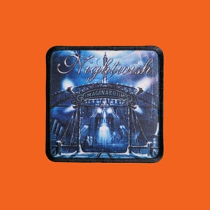 Könnte beinhalten: Ein quadratischer Aufnäher mit schwarzem Rand mit dem Bandnamen "Nightwish" in silberner Schrift über einem Bild einer Bühne mit dem Wort "IMAGINAERUM" in Silber. Der Hintergrund ist blau.