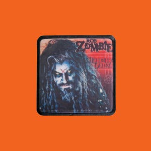 Peut inclure: Un écusson carré avec un portrait de Rob Zombie aux longs cheveux noirs et maquillage. L'écusson porte les inscriptions "Rob Zombie" et "Hellbilly Deluxe" en lettres rouges et blanches.