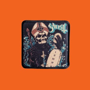 Op de afbeelding: Vierkante zwarte patch met een skeletfiguur in een bisschopshoed, die een tablet met Romeinse cijfers vasthoudt. Het woord "Ghost" staat in blauwe letters. Het ontwerp bevat blauwe, waterachtige elementen.