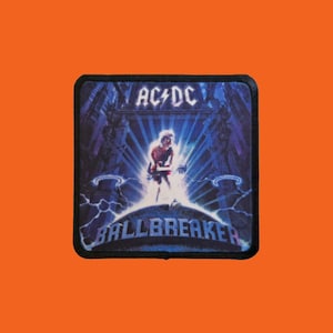 Peut inclure: Écusson carré avec le logo AC/DC au-dessus du mot "BALLBREAKER". L'image montre un guitariste sur scène avec un fond bleu et violet. L'écusson a une bordure noire.