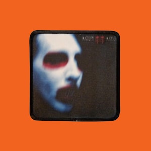 Peut inclure: Écusson carré avec un portrait d'une personne à la peau blanche, maquillage rouge pour les yeux et bouche ouverte. Le nom "Marilyn Manson" est imprimé en rouge en haut à droite. L'écusson a une bordure noire et est posé sur un fond orange.