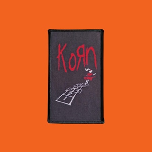 May include: Black rectangular patch with the red word "Korn" in a stylized font. A hopscotch design with numbers 1 to 4 is below the word. A small figure is jumping on the hopscotch. The patch has a black border.
