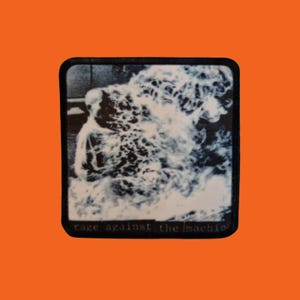 May include: A black and white patch featuring the iconic image of a Buddhist monk on fire. The text "rage against the machine" is printed in white at the bottom of the patch. The patch has a black border and is set against an orange background.