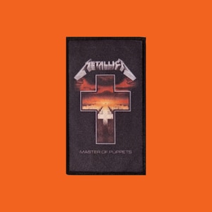 Può includere: Una toppa rettangolare nera con il logo Metallica in argento sopra un disegno a croce con uno sfondo di tramonto. Le parole "MASTER OF PUPPETS" sono stampate sotto in bianco.