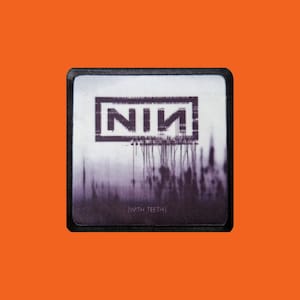 May include: A square patch with a black border featuring the band name "NIN" in a rectangular frame. Below the frame, a blurred purple and white design with the text "(WITH TEETH)" is visible. The patch is set against an orange background.