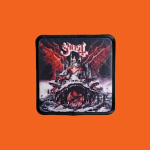 Könnte beinhalten: Quadratischer schwarzer Aufnäher mit dem Bandnamen "Ghost" in weißen gotischen Buchstaben. Der Aufnäher zeigt eine detaillierte, düstere Fantasieszene mit einer Burg, geflügelten Kreaturen und einer feurigen, höllischen Landschaft in Rot- und Schwarztönen.
