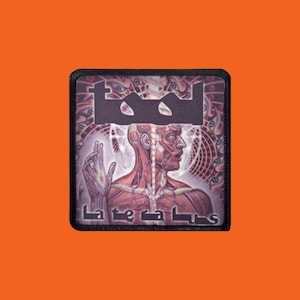 Peut inclure: Écusson carré à bordure noire avec le nom du groupe "Tool" et le titre de l'album "Lateralus". Le motif comprend une illustration anatomique détaillée d'une figure humaine avec un arrière-plan complexe.