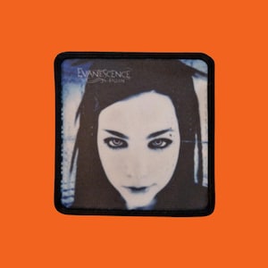 Peut inclure: Écusson carré avec une bordure noire, représentant un visage féminin en noir et blanc. Le texte "Evanescence" et "Fallen" est imprimé en blanc sur un fond bleu.