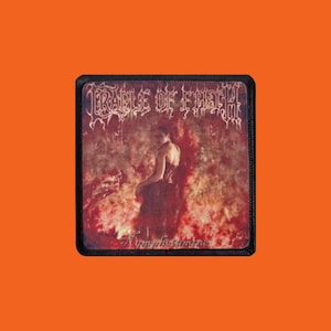 Op de afbeelding: Vierkante patch met een zwarte rand met de bandnaam "Cradle of Filth" in sierlijke letters boven een afbeelding van een persoon in een donkere jurk. Het woord "Nymphetamine" staat in cursief onderaan. De achtergrond is een mix van rood en bruin.