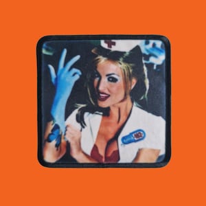 Può includere: Toppa quadrata raffigurante una donna in uniforme da infermiera, con cappello bianco e croce rossa. Indossa un reggiseno rosso e un guanto blu, con un tatuaggio a farfalla. La toppa ha un bordo nero e la scritta "Blink 182".