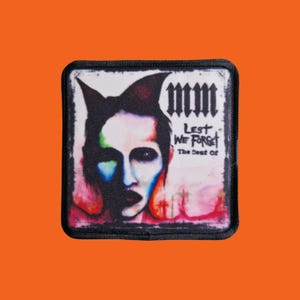 May include: A square patch featuring a portrait with a black and white color scheme, with accents of red, blue, and green. The text "mm" is at the top, with "Lest We Forget" and "The Best Of" below the portrait. The patch has a black border.