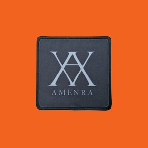 Peut inclure: Un écusson carré gris foncé avec une bordure noire. L'écusson présente un symbole double-A blanc stylisé au-dessus du mot "AMENRA" en blanc, centré sur l'écusson.