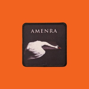 May include: A black square patch featuring the word "AMENRA" in white capital letters. Below, a white, stylized swan-like figure with a red hand. The patch has a black border and is set against an orange background.