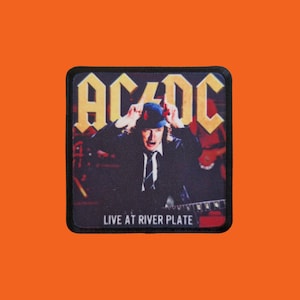May include: A square patch featuring the band AC/DC with the band's logo in yellow. The image shows a live performance with the text "LIVE AT RIVER PLATE" at the bottom. The patch has a black border and is set against an orange background.