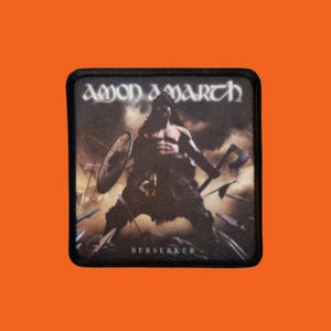 Puede incluir: Un parche cuadrado negro con el nombre de la banda "Amon Amarth" en letras blancas sobre una ilustración de un guerrero. El guerrero sostiene un hacha y un escudo. La palabra "Berserker" está impresa en blanco en la parte inferior.