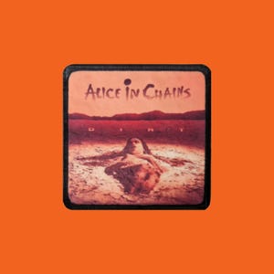 Peut inclure: Écusson carré avec la pochette de l'album "Dirt" d'Alice in Chains. L'image montre une figure immergée dans un paysage brun-rougeâtre, avec le nom du groupe et le titre de l'album en haut.