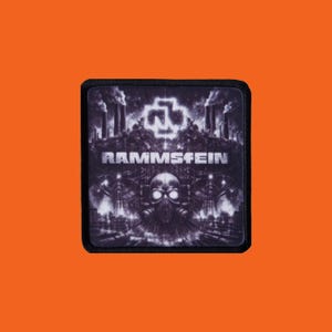 Peut inclure: Un écusson carré à bordure noire. Le design en niveaux de gris présente le nom du groupe "RAMMSTEIN" en blanc, un logo stylisé et un crâne portant un masque à gaz. L'arrière-plan représente des structures industrielles.